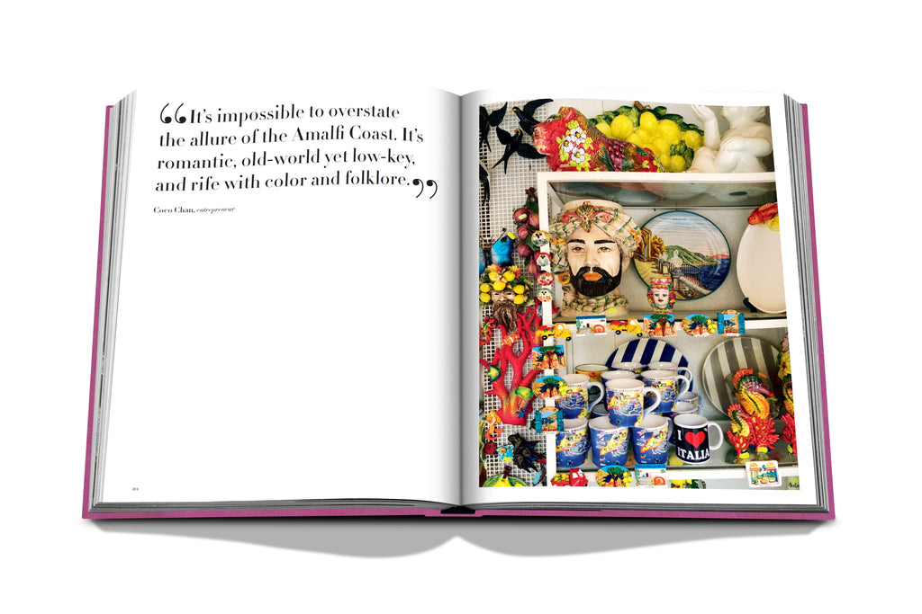An open copy of "Amalfi Coast" by Assouline displays a quote about the côte Amalfitaine on the left page, while the right page showcases a colorful assortment of ceramic mugs, bowls, and vases. A decorative figure's head is elegantly surrounded by lemons and vibrant items.