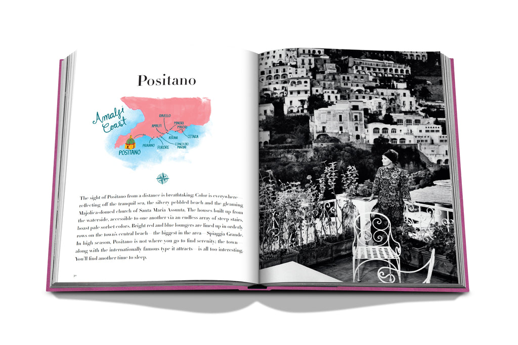 The product, *Amalfi Coast* by Assouline, features an open page showcasing a colorful map of the Côte Amalfitaine on the left. On the right, there's a black-and-white photograph of a woman standing by a railing, gazing at Positano's hillside structures and pebble-lined beaches below.