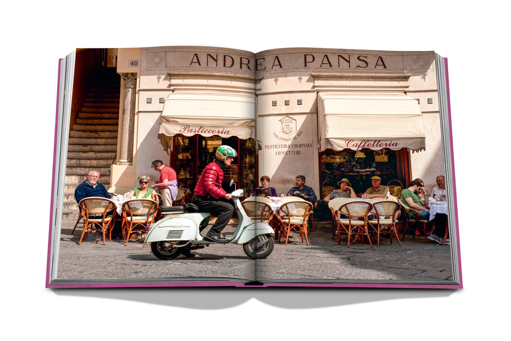 A person rides a scooter past a café with outdoor seating, capturing the quaint charm of the Amalfi Coast. Several patrons enjoy their time at tables beneath awnings that read Pasticceria Confetteria and Caffettoria, evoking images of scenic views and the allure of Assouline's panoramiques randonnées nearby.